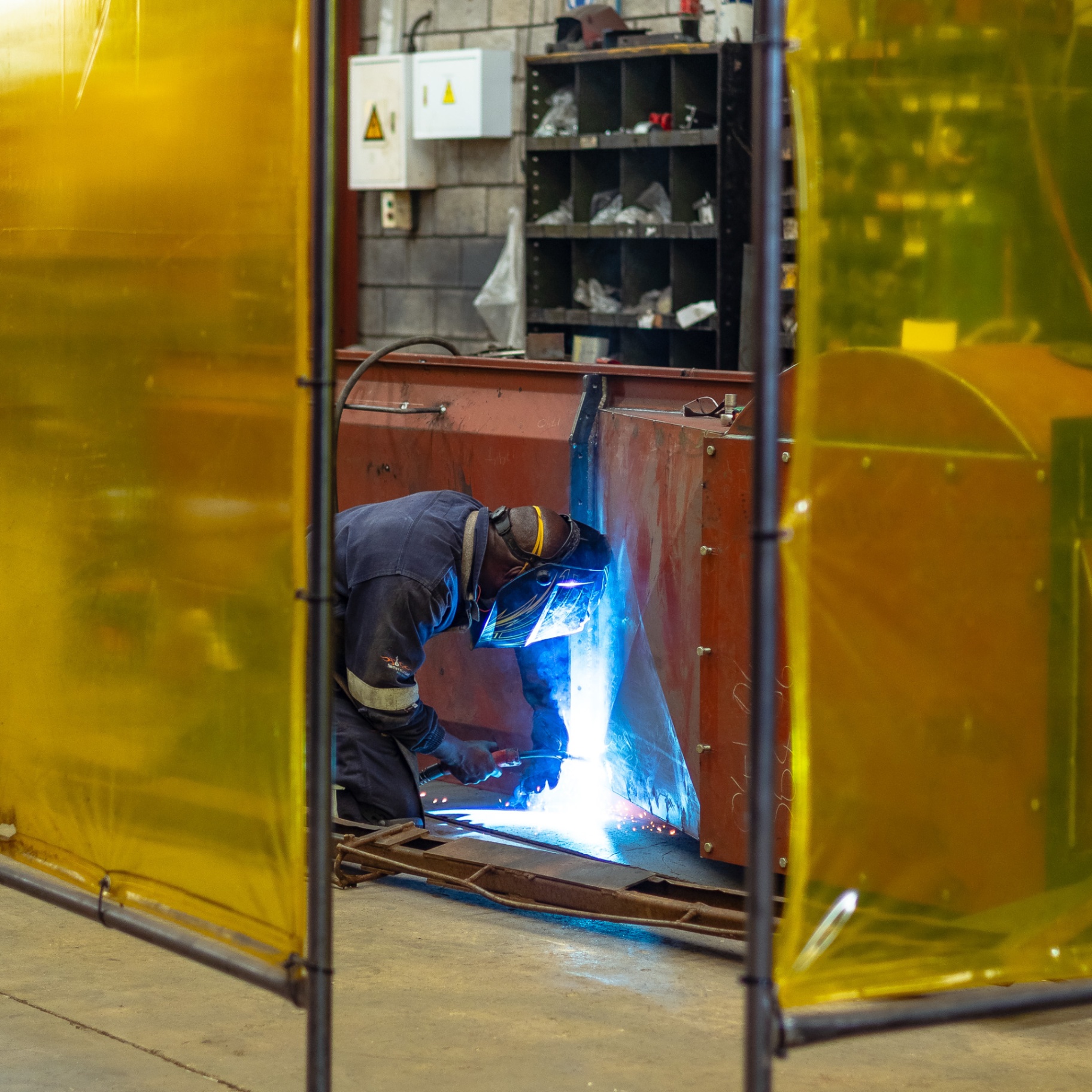 Welder working on a steel panel behind protective yellow welding curtains